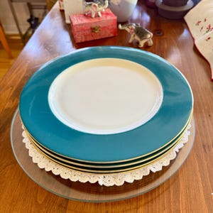 Può includere: Una pila di piatti in ceramica turchese e bianchi con bordatura dorata, appoggiati su un piatto di vetro trasparente e un centrino di pizzo color crema. I piatti hanno un bordo turchese e un centro bianco.