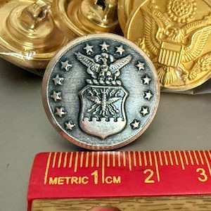 May include: A collection of military uniform buttons. One button is silver-toned with the Air Force emblem, surrounded by stars. Other buttons are gold-toned with an eagle emblem. A red metric ruler is in the foreground.