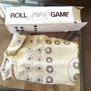 May include: A Roll Game board game set featuring a rectangular white cardboard box with the text ROLL GAME printed in brown, alongside a beige fabric game mat printed with a repeating brown circular pattern that matches the box design.