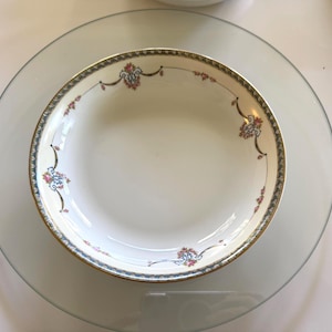 Puede incluir: Un cuenco de porcelana blanca con borde dorado y diseño floral, colocado sobre un plato de cristal transparente. El cuenco tiene un borde decorativo con detalles azules y rojos. El plato de cristal es redondo.
