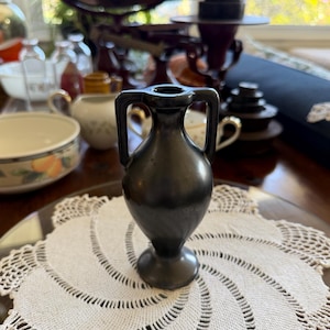 May include: A small, matte black ceramic amphora vase featuring a narrow neck, a flared circular base, and two symmetrical vertical handles extending from the rim to the shoulder of the vessel, standing upright on a white crocheted doily.