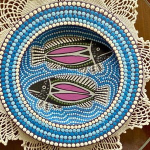 May include: A decorative, hand-painted bowl featuring two stylized fish. The bowl's rim is adorned with blue dots, surrounding a central design of fish with black, white, and pink details. The bowl is placed on a white lace doily.