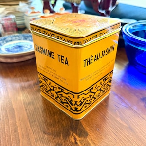May include: A square, yellow Jasmine Tea tin with black text and decorative patterns. The tin reads "Jasmine Tea" and "The Au Jasmin". The product is from The People's Republic of China.