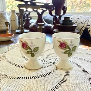 May include: Two white porcelain egg cups featuring a delicate pink rosebud design with green leaves and a thin gold rim. Each cup has a rounded bowl shape supported by a sturdy pedestal base, crafted from smooth, glossy ceramic material.