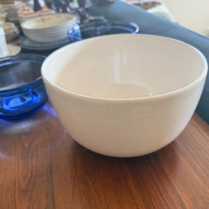 May include: A large, white ceramic bowl with a rounded shape sits on a wooden surface. A smaller, blue glass bowl is visible to the left. Other dishware is in the background.