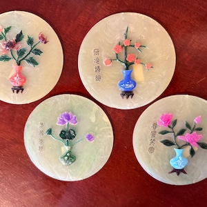 May include: Four round, off-white coasters with floral designs. Each coaster features a different flower arrangement in a small vase. The vases are in various colors, including red, blue, and green. The coasters have Chinese characters.