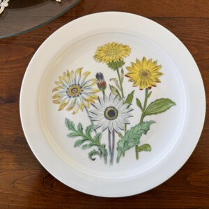 May include: White ceramic plate featuring a floral design with yellow and white flowers, green leaves, and stems. The plate has a wide rim and is set against a dark wood surface. The flowers are detailed and realistic.