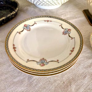 Puede incluir: Una pila de tres platos de porcelana blanca con borde dorado y un diseño floral y de volutas decorativo en rosa, azul y dorado. Los platos están sobre un mantel de lino de color neutro.
