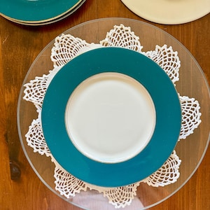 May include: Overhead view of a teal and white dinner plate set on a clear glass charger plate with a white lace doily. Additional plates with teal rims are visible in the background. The plates are arranged on a wooden surface.