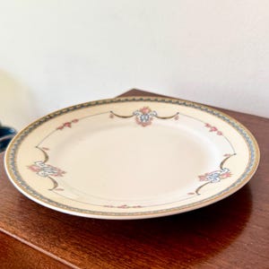 Puede incluir: Un plato ovalado de color crema con un borde decorativo. El borde presenta un diseño azul y dorado con detalles florales en rosa y azul. El plato está sobre una superficie de madera oscura.