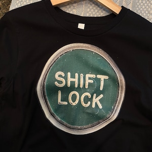 May include: Black t-shirt featuring a green and white graphic of a vintage-style key with the text "SHIFT LOCK" printed on it.