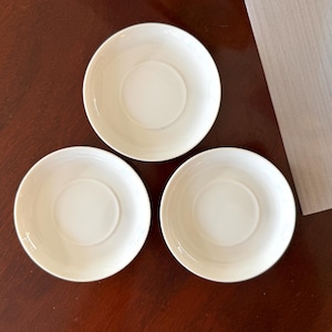 May include: Three circular white ceramic saucers arranged in a triangular formation. Each saucer features a smooth, solid white surface with a slightly raised central circular indentation designed to hold a cup securely in place.
