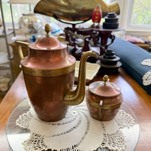 May include: A hammered copper coffee pot and a matching sugar bowl, both featuring brass accents, a decorative leaf-patterned band, and small brass finials on their lids. The coffee pot has a curved spout and a handle, while the sugar bowl has two handles.