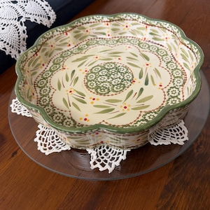 Pode incluir: Um prato de cerâmica redondo com borda recortada, apresentando uma base de cor creme decorada com padrões de folhas verdes, pequenos pontos amarelos e vermelhos, e um motivo circular central composto por intrincados desenhos florais verdes e brancos.