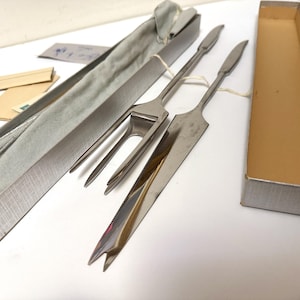 May include: A set of stainless steel serving utensils, including a carving fork and a serving knife, presented in a silver box with a gray lining. The set is accompanied by an empty box and small cards.