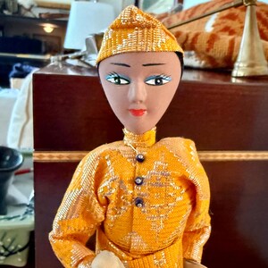 May include: A vintage doll with a painted face, wearing a gold sequined outfit and matching hat. The doll has blue eye shadow, red lips, and a high collar. The doll's outfit has dark blue buttons.