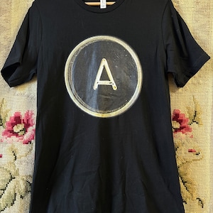 May include: Black t-shirt featuring a large, circular graphic of a typewriter key with the letter "A" in the center. The shirt is hanging on a wooden hanger against a patterned background. The shirt is made of a soft material.
