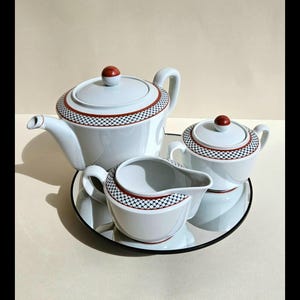 Puede incluir: Un juego de té de porcelana blanca con un patrón geométrico rojo y negro. El juego incluye una tetera, una jarra de crema, un azucarero y una bandeja de espejo.