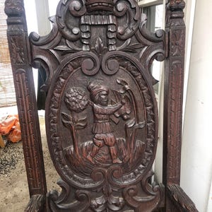 Può includere: Sedia antica in legno marrone scuro con intagli intricati. Lo schienale della sedia presenta un'intagliatura a rilievo dettagliata di una figura in un paesaggio, incorniciata da volute ornate e sormontata da una corona. Anche i braccioli e le gambe sono intagliati.