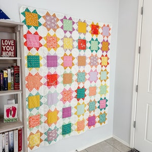 May include: A colorful quilt with a star pattern in shades of pink, purple, yellow, green, and orange. The quilt is hanging on a white wall.