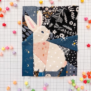 Może przedstawiać: Patchworkowy królik z tkaniny, z różowymi uszami i jasnoróżowym i białym korpusem. Królik jest osadzony na tle granatowej i czarnej tkaniny kwiatowej. Kolorowe gwiazdki origami są rozrzucone wokół królika.