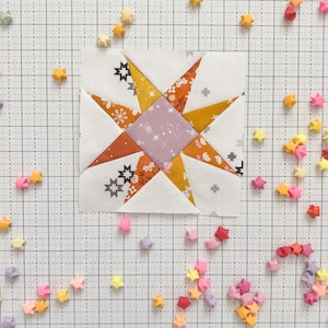 May include: A patchwork quilt block featuring a star design in shades of orange, yellow, purple, and white. The block is surrounded by small origami stars in various colours.