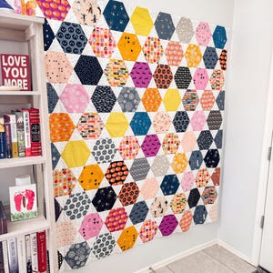May include: A large, colourful quilt with a honeycomb pattern, featuring various Halloween-themed fabrics. The quilt is primarily white, with hexagonal patches in orange, black, pink, and blue, adorned with pumpkins, bats, and other festive designs. The quilt is hanging on a white wall.