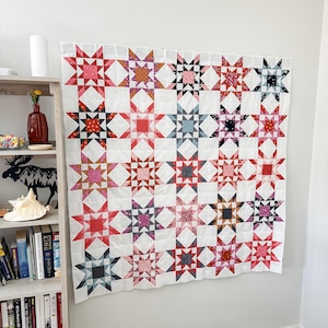 Op de afbeelding: Een vierkante patchwork quilt met een herhalend sterpatroon in diverse kleuren, waaronder rood, roze, blauw en oranje, tegen een witte achtergrond. De quilt is samengesteld uit meerdere stoffen blokken in een raster, met diverse bloemen- en geometrische prints in elk sterontwerp.