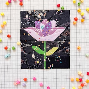 Może przedstawiać: Blok kwiatowy z tkaniny z czarnym tłem, przedstawiający fioletowy i różowy kwiat z zieloną łodygą. Kwiat otoczony jest kolorowymi gwiazdami origami. Blok znajduje się na białej siatce.