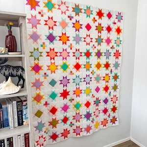 May include: A large square quilt featuring a repeating star pattern in vibrant colors including pink, orange, yellow, red, purple, and teal. The stars are set against a white background with small colorful square accents between the star blocks.