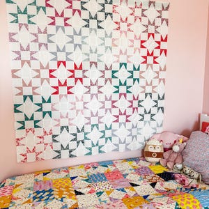 May include: A large, colorful quilt with a star pattern hangs on a pink wall. The quilt features a variety of fabrics in shades of red, green, and blue. A smaller quilt with a patchwork design covers the bed. Plush toys are on the bed.