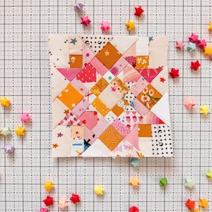Może przedstawiać: Kwadratowy blok patchworkowy z geometrycznym wzorem w odcieniach różu, pomarańczy i bieli. Blok otoczony jest kolorowymi gwiazdami origami rozrzuconymi na tle kratki. Blok patchworkowy zawiera różne wzory.