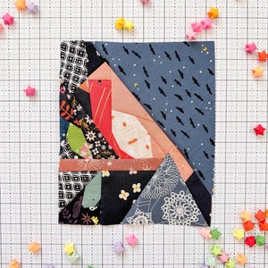 Może przedstawiać: Kwadratowy blok patchworkowy z różnymi wzorami tkanin w odcieniach niebieskiego, różowego, czerwonego i czarnego. Blok otoczony jest kolorowymi gwiazdami origami, rozrzuconymi na tle kratki.