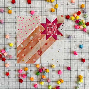 Może przedstawiać: Ręcznie wykonany blok do pikowania z różowym wzorem gwiazdy i różnymi wzorzystymi tkaninami. Blok otoczony jest kolorowymi gwiazdami origami rozrzuconymi na tle kratki, tworząc zabawną i artystyczną kompozycję.