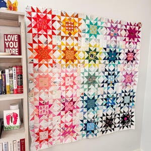 Puede incluir: Una colcha colorida con un patrón de estrellas. La colcha está hecha de muchas telas y colores diferentes, incluyendo rojo, naranja, amarillo, verde, azul, morado y blanco. La colcha está colgada en una pared blanca.