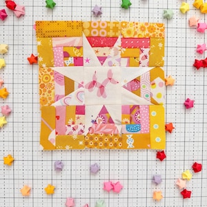 Może przedstawiać: Kolorowy blok patchworkowy z motywem gwiazdy, z różnymi wzorami i odcieniami żółtego, różowego i białego. Origami gwiazdy w wielu kolorach są rozrzucone wokół bloku na tle kratki.