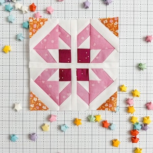 Puede incluir: Un bloque de colcha de retazos rosa y blanco con un borde floral. El bloque está rodeado de estrellas de origami de colores sobre una superficie blanca cuadriculada.