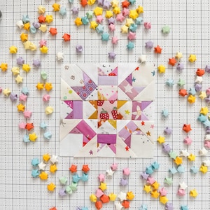 Może przedstawiać: Kolorowy blok patchworkowy z motywem gwiazdy, otoczony rozrzuconymi gwiazdami origami w różnych pastelowych kolorach. Blok patchworkowy zawiera różowe, fioletowe i żółte tkaniny ze wzorami truskawek i gwiazd. Tło to kratka.