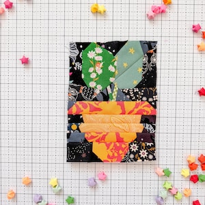 Może przedstawiać: Kolorowe dzieło sztuki z tkaniny przedstawiające stylizowaną roślinę. Wykorzystuje tkaniny we wzory w kolorze zielonym, pomarańczowym i czarnym. Obraz zawiera tło w kratkę i rozrzucone gwiazdy origami w różnych kolorach. Projekt przypomina blok patchworku.