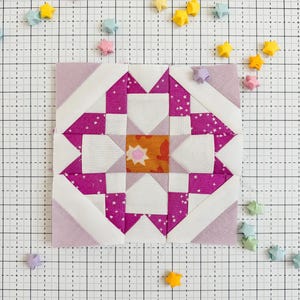 May include: A pink and white patchwork quilt block with a star design. The block is made up of squares and triangles of different fabrics. The block is surrounded by a grid of small squares.