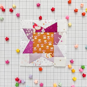 May include: A patchwork quilt block with a star design in shades of pink, purple, orange, and white. The block is surrounded by small origami stars in various colors on a white grid background.