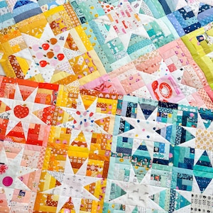 May include: A patchwork quilt with a star design in a variety of colours. The quilt features a mix of fabrics in shades of blue, yellow, orange, and pink. Some blocks have star designs, while others have geometric patterns and small illustrations. The quilt is a mix of textures and colours.