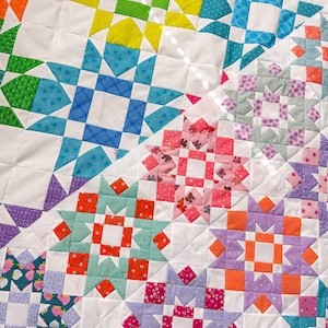 May include: Close-up of a colorful quilt with a star pattern. The quilt features a variety of colors, including orange, yellow, green, blue, pink, and purple, set against a white background. The quilt is made of squares and triangles.