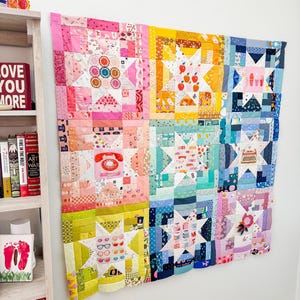 Może przedstawiać: Kolorowa patchworkowa kołdra z różnorodnymi wzorami i wzorami, w tym gwiazdami, jabłkami i kształtami geometrycznymi. Kołdra wykonana jest z różnych tkanin w odcieniach różu, żółci, niebieskiego i zieleni. Kołdra wisi na białej ścianie.