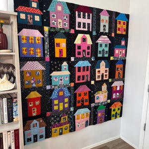 May include: A large rectangular quilt featuring a grid of twenty-five colorful fabric houses against a black background. Each house has a unique roof color and pattern, with windows displaying various prints including pumpkins, cats, and stars.