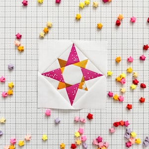 May include: A pink and gold star-shaped quilt block with a white background. The block is surrounded by small origami stars in various colors, including red, orange, yellow, green, blue, purple, and pink.