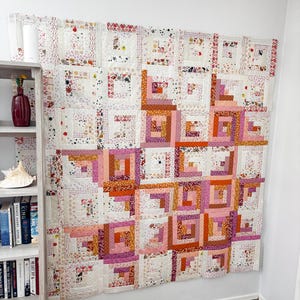 May include: A large, handmade quilt with a log cabin design, displayed on a white wall. The quilt features squares of various pink, orange, and white patterned fabrics. The quilt is a patchwork of squares, creating a warm and inviting aesthetic.