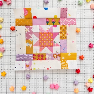 Może przedstawiać: Kolorowy blok patchworkowy z motywem gwiazdy, z różowych, żółtych i białych tkanin. Blok otoczony jest gwiazdami origami w różnych kolorach. Blok ma wymiary około 25 cm kwadratowych.