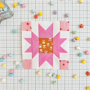 May include: A pink and white patchwork quilt block with a star design. The block is surrounded by small origami stars in various colors.