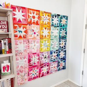 Peut inclure: Une grande courtepointe colorée avec un motif d'étoiles, exposée sur un mur blanc. La courtepointe présente un dégradé arc-en-ciel de couleurs, du rouge au bleu, avec des motifs d'étoiles blanches. La courtepointe est un patchwork de divers tissus et motifs.